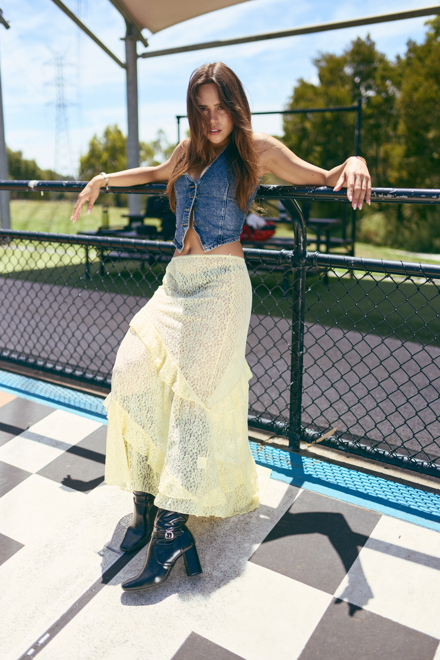 Young woman in denim vest and lace skirt leans against a fence outdoors on a sunny day, wearing black boots.  